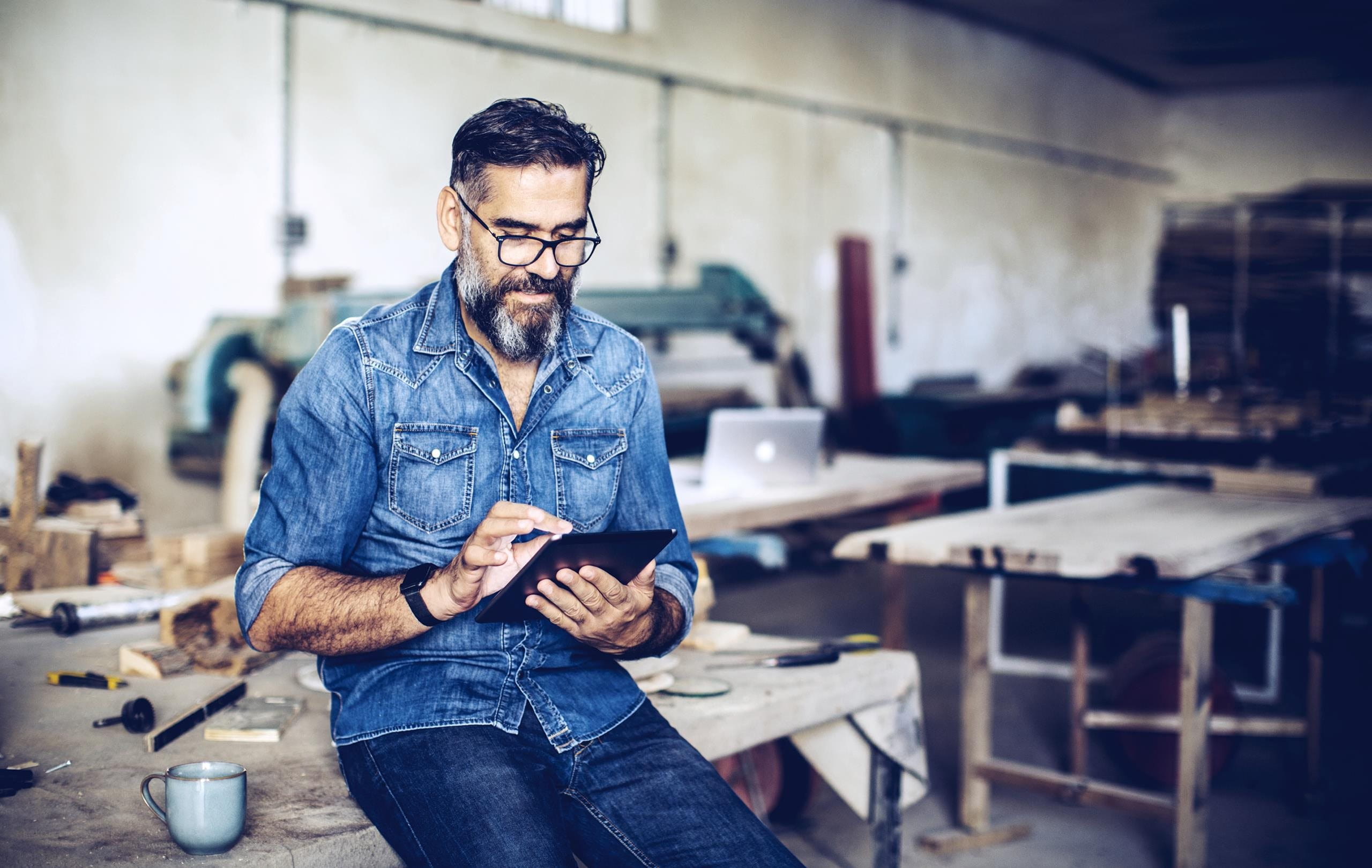 Mann sitzt auf Werkbank und schaut ins iPad Mann sitzt auf Werkbank und schaut ins iPad